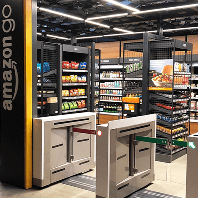 Automated exit and entrance of Amazon Go, a self-serve grocery store. Image shows black shelving with food items and a subway-style automated entrance and exit bar.
