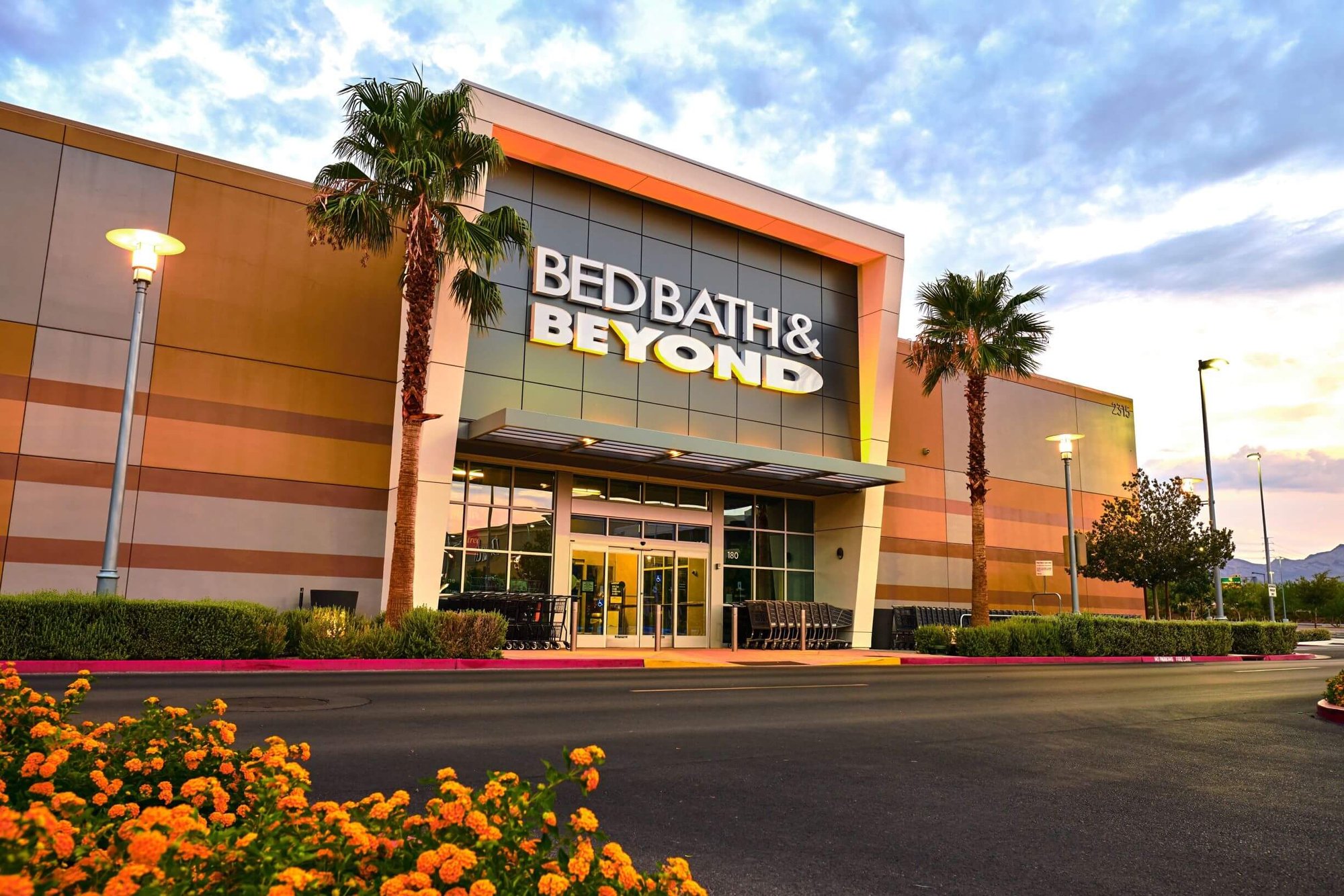 Bed Bath & Beyond storefront at dusk, with palm trees at the entrance and lights in the entryway
