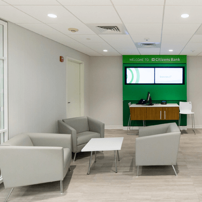 Three grey, oversized chairs and a white reception table inside Citizen's bank, with a green accent wall reading 'Welcome to Citizens Bank'.