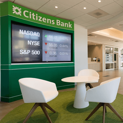 Three white chairs and a table inside Citizen's bank, looking at a large LCD screen showing the latest stock market updates.