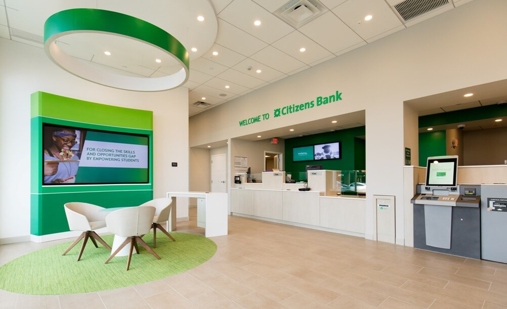 Interior lobby of Citizen's Bank Charlestown, showing bright white interiors with green accents. A large sign reads 'Welcome to Citizens Bank'.