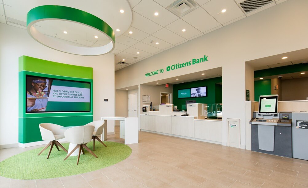 Interior entrance of Citizen's Bank Charlestown, showing bright white interiors with green accents. A large sign reads 'Welcome to Citizens Bank'.