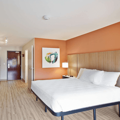 Guest room in an Extended Stay America hotel, featuring a large bed with white linens and bright orange walls.