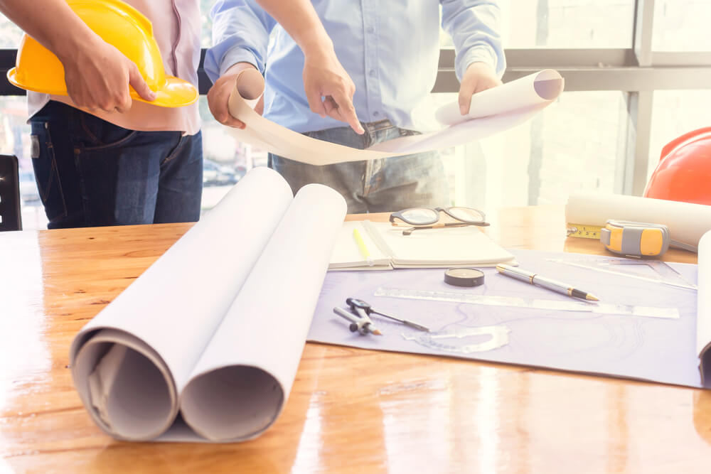 Man with yellow hard hat and businessman pointing to architectural drawings at a construction site.