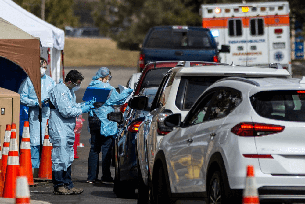 Line of vehicles with medical professionals standing in blue protective gear, showing Covid-19 testing facility.