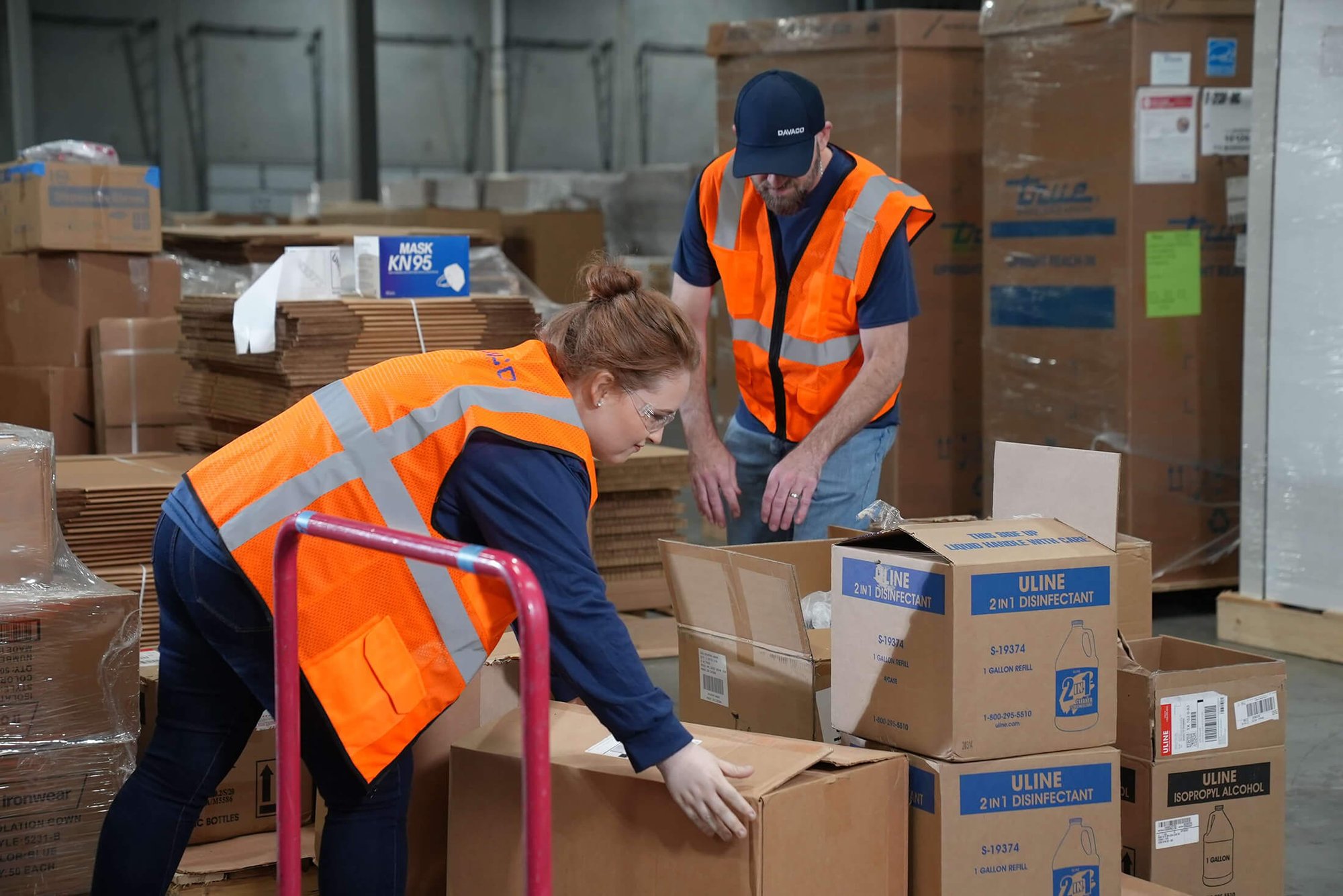 Two DAVACO logistics employees wearing orange safety vests working with boxes in a warehouse, showing logistics service.