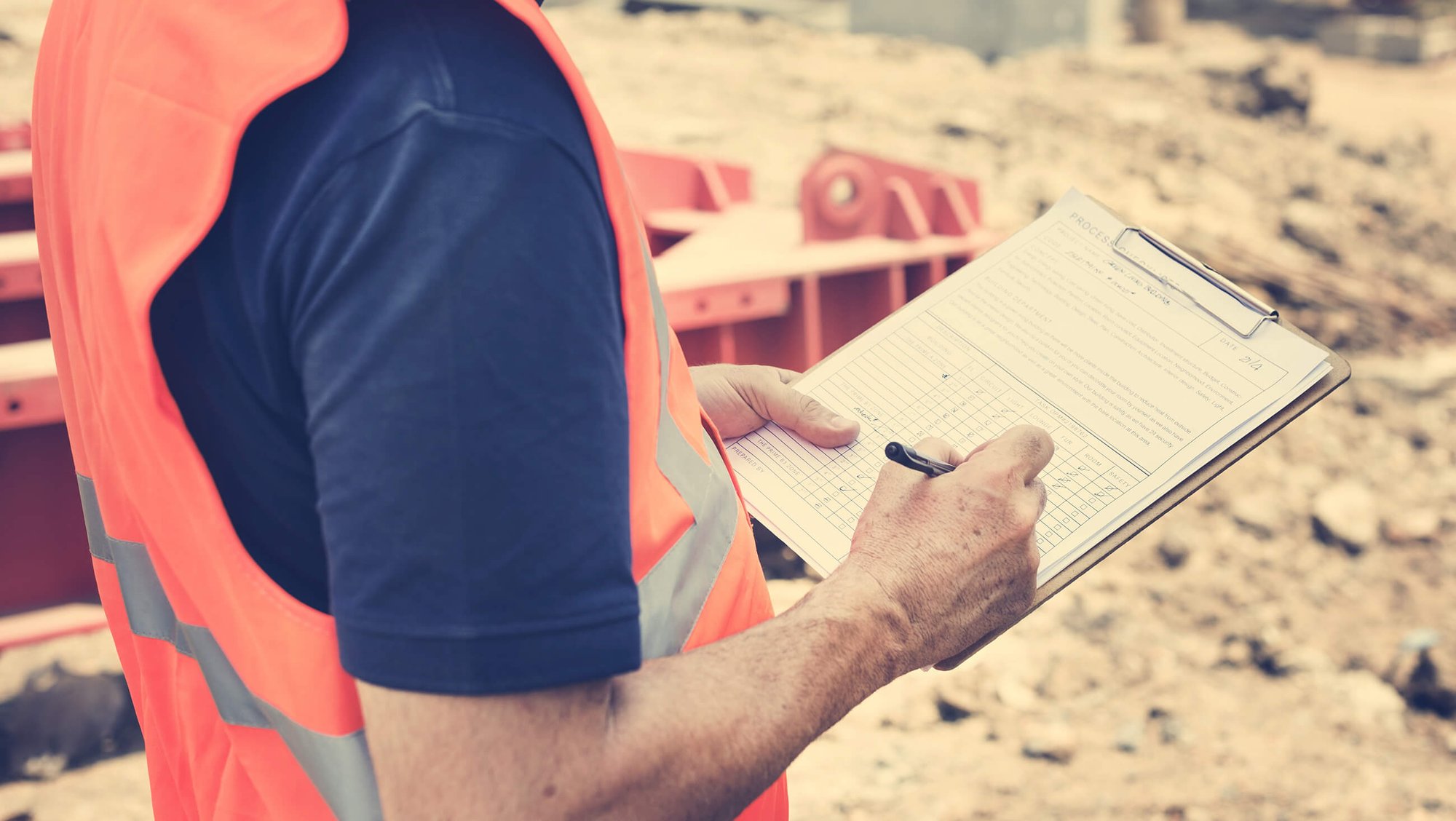 DAVACO employee wearing a blue shirt and orange safety vest, holding a clipboard and pen at an exterior construction site.
