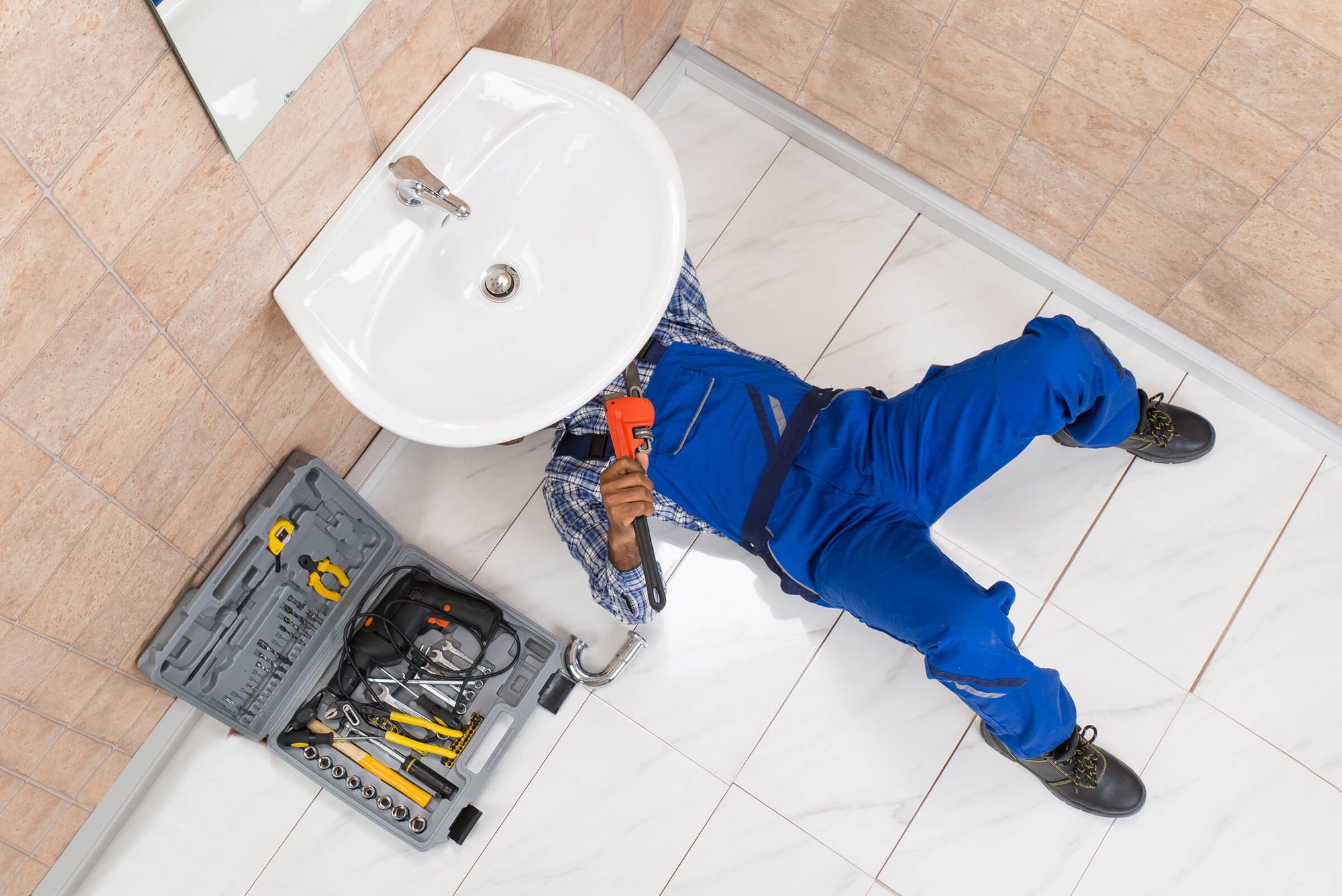 DAVACO plumber, wearing a bright blue uniform, working underneath a wall-mounted sink in a customer's bathroom.