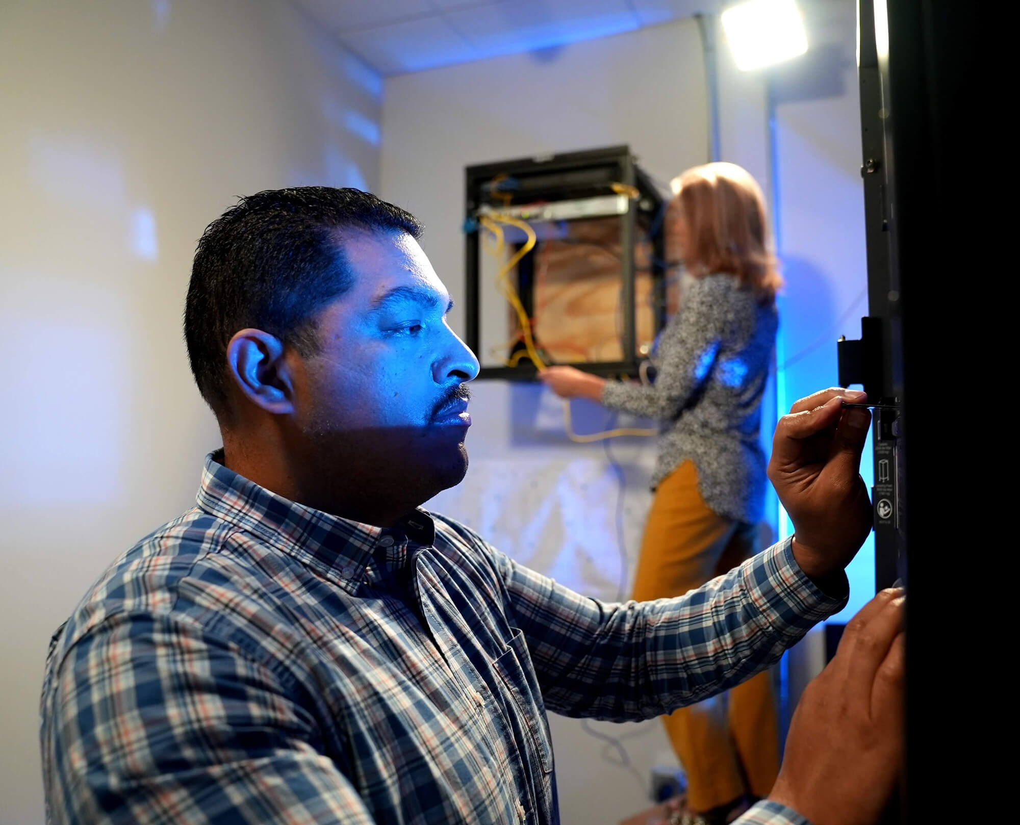 Male DAVACO employee wearing a plaid button shirt working on equipment in a technology storage room.