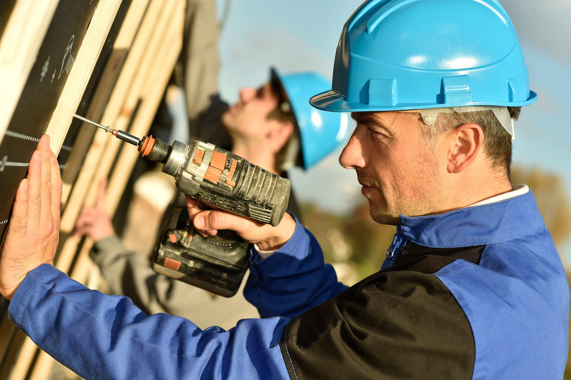 Male construction worker wearing a blue hard hat and vest drilling into wood at an exterior construction site.
