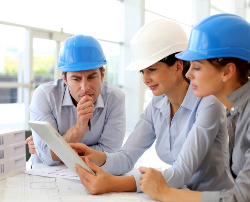 Three employees in business shirts wearing blue and white hard hats studying a digital notebook with program plans.