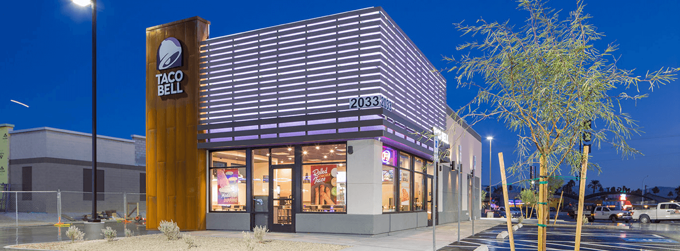 Night view of a Taco Bell restaurant, purple lights illuminating the storefront, with colorful signs in the windows advertising new menu items.